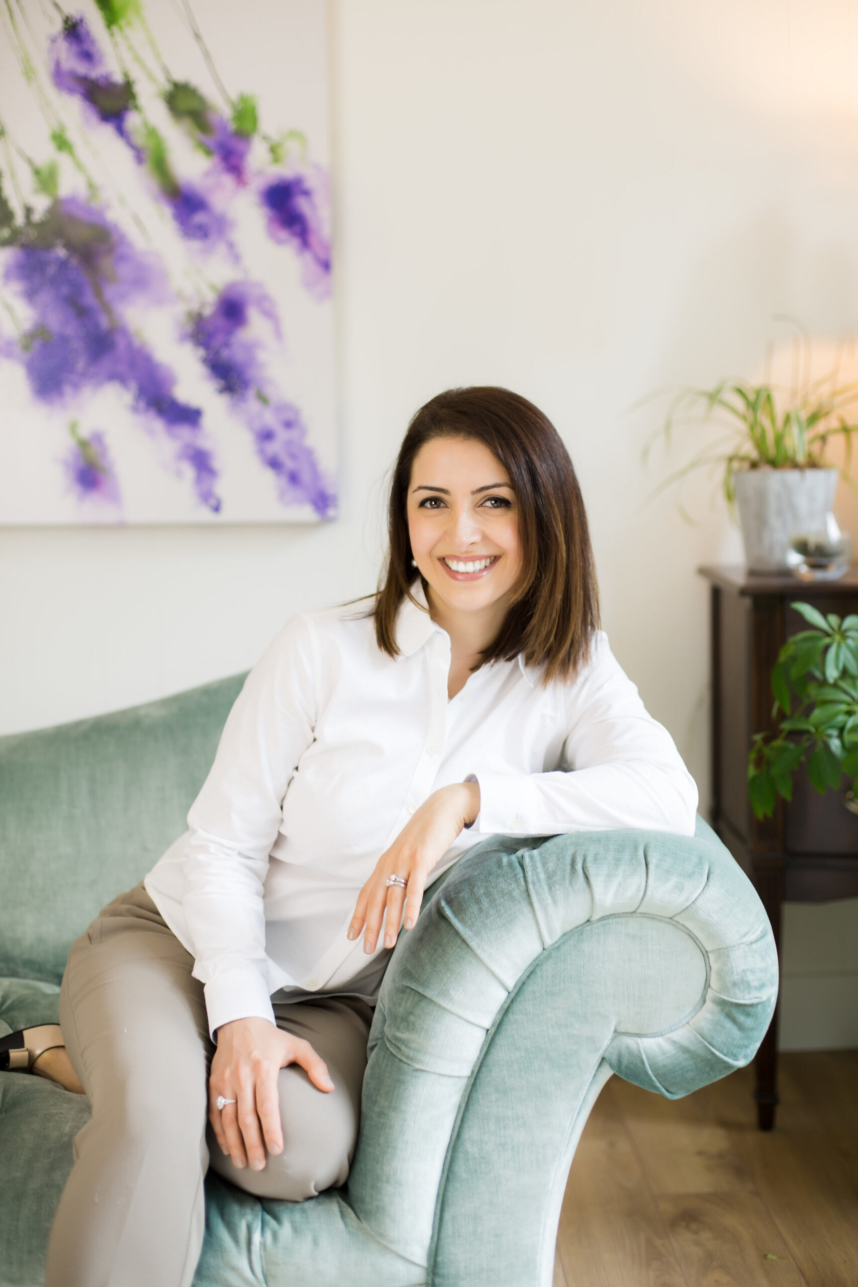 Dr. Mona Afrand leaning on a mint green couch smiling with some plants in the background.