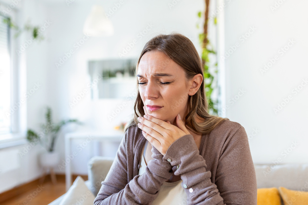 Woman siting on a couch holding her jaw with both hands in pain.