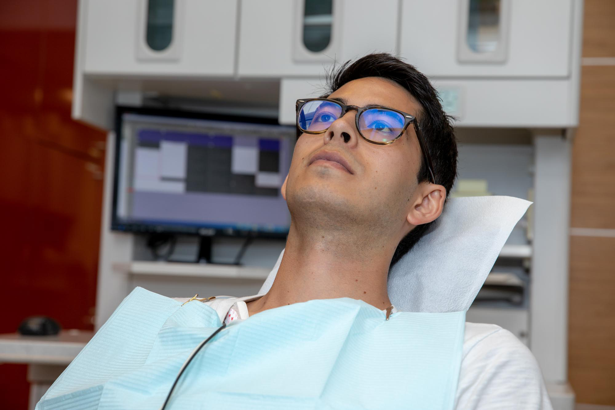 Man with glasses reclined in a dentist chair wearing a dental bib with a dental equipment behind them.